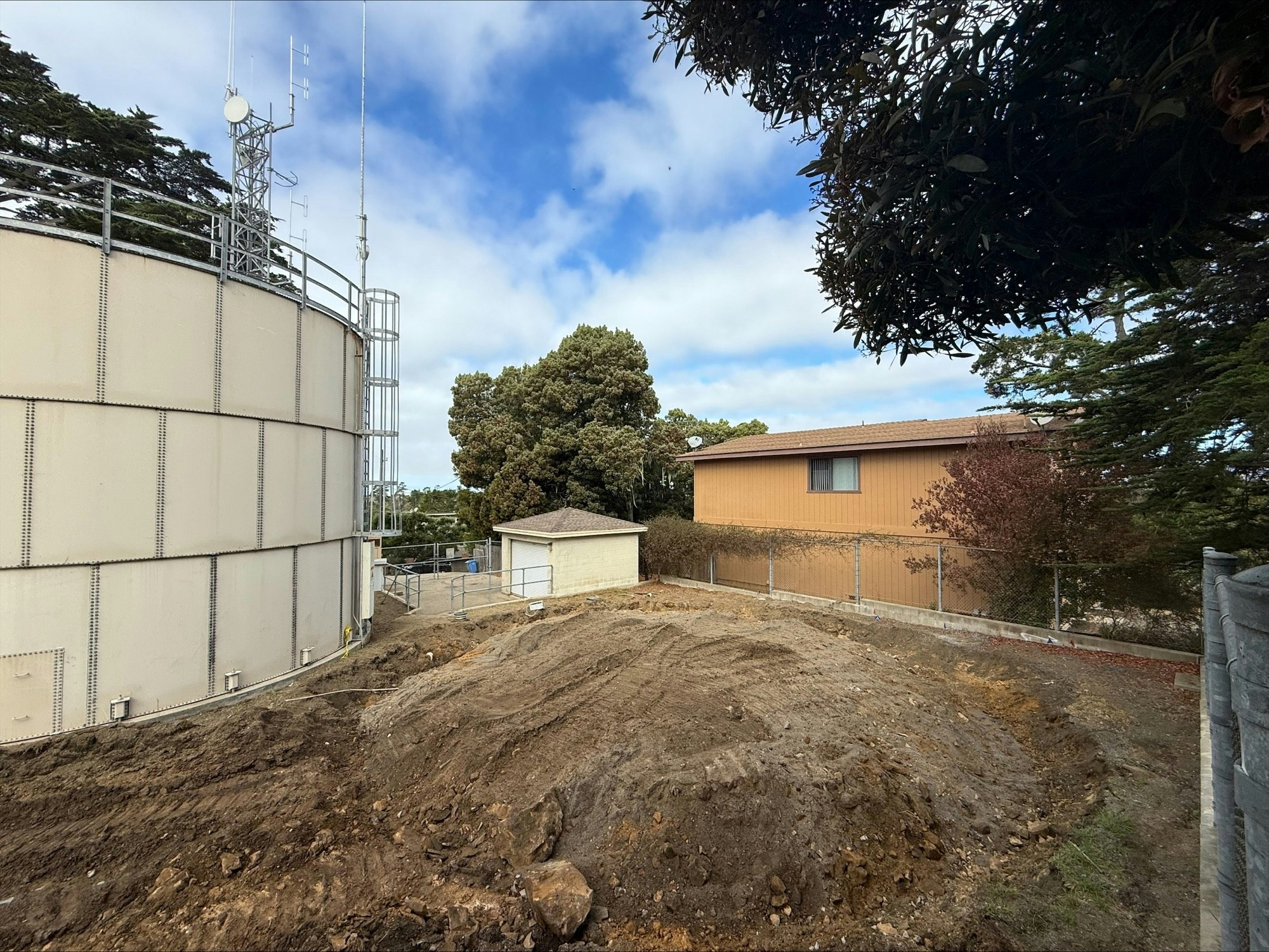 Large water tank, dirt mound, small building, and a house surrounded by trees and a fence under a partly cloudy sky.