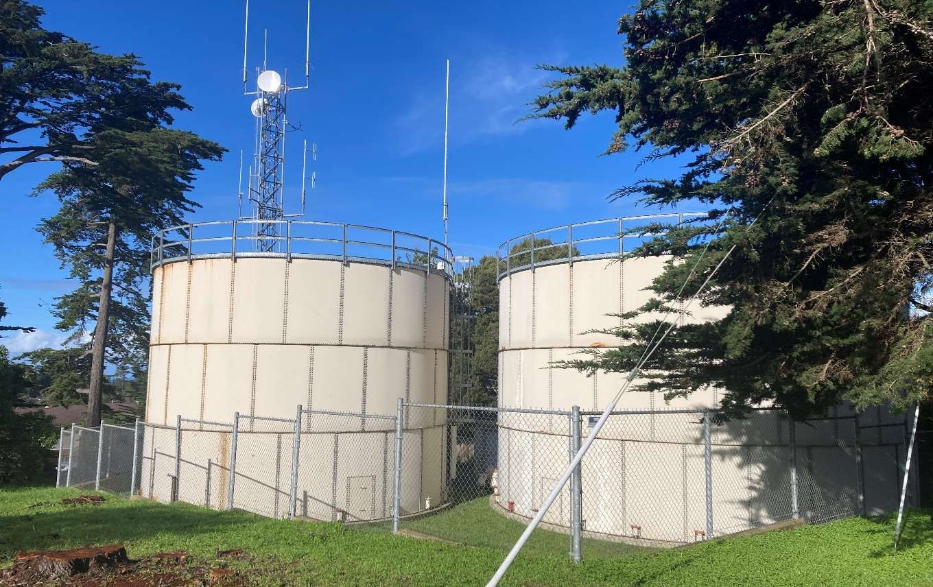 Two large cylindrical tanks fenced off with antennas on top, surrounded by trees and grass.