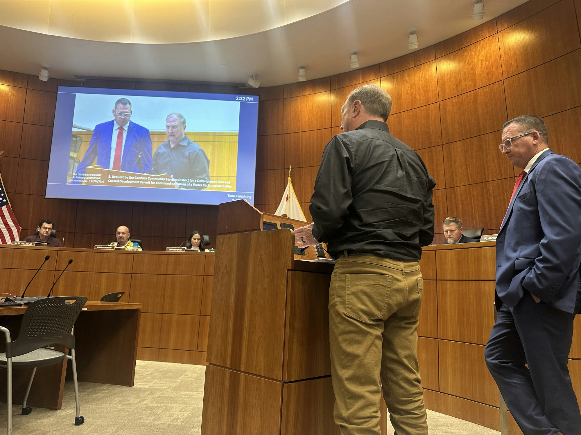 A public meeting with officials discussing a development project, featuring presenters and a screen showing details.