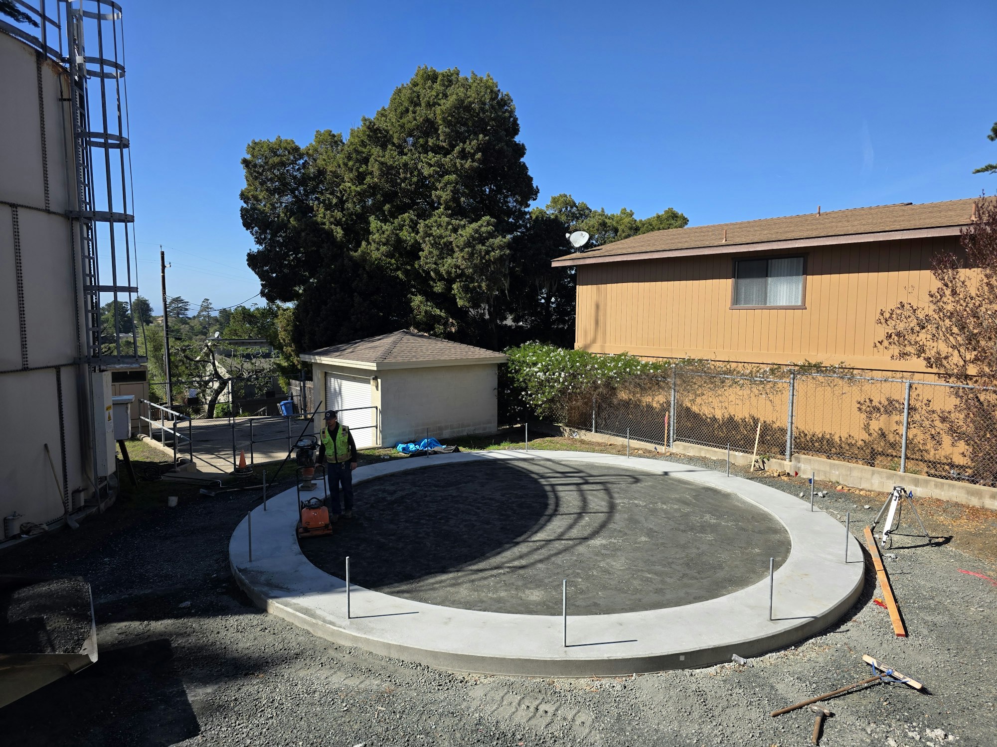 Construction site with a circular foundation, worker with equipment, trees, a brown building, and clear blue sky.