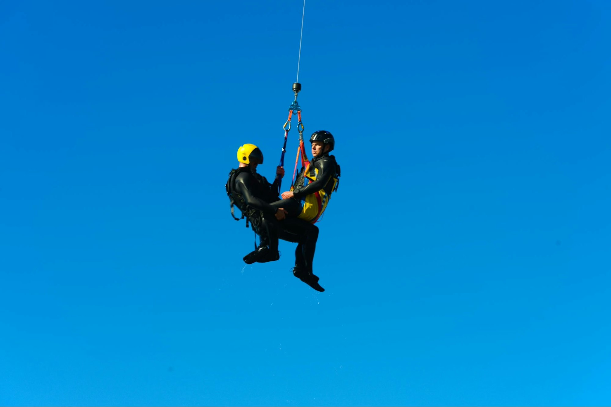 Two individuals are being airlifted, one in a yellow helmet, suspended from a cable against a clear blue sky.