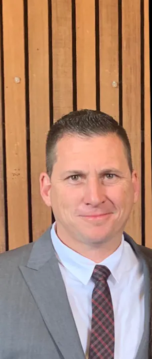 A man in a suit with a tie is smiling, standing in front of a wooden-paneled background. He appears professional and approachable.