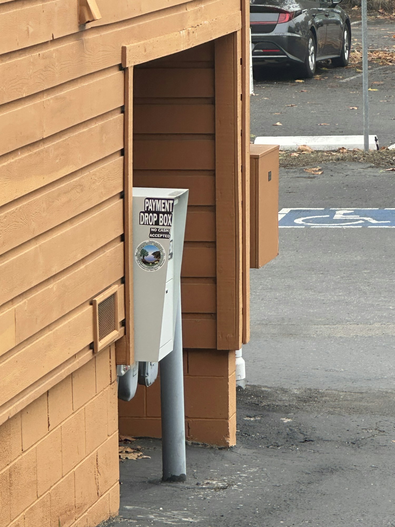 Payment drop box at a wooden structure, with a parked car and handicap parking sign nearby.