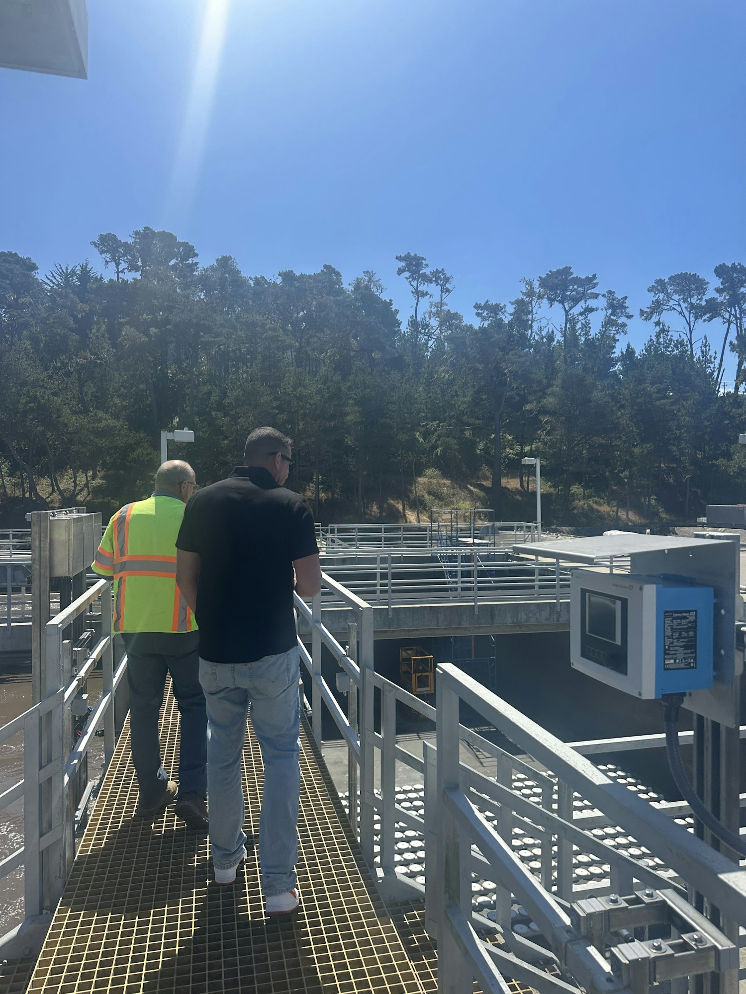 Two people on a metal walkway with railing, one in a high-visibility vest, near water treatment or industrial facility. Trees in the background.