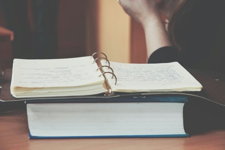 A person is sitting next to an open ring binder with handwritten notes, placed on a thick book.