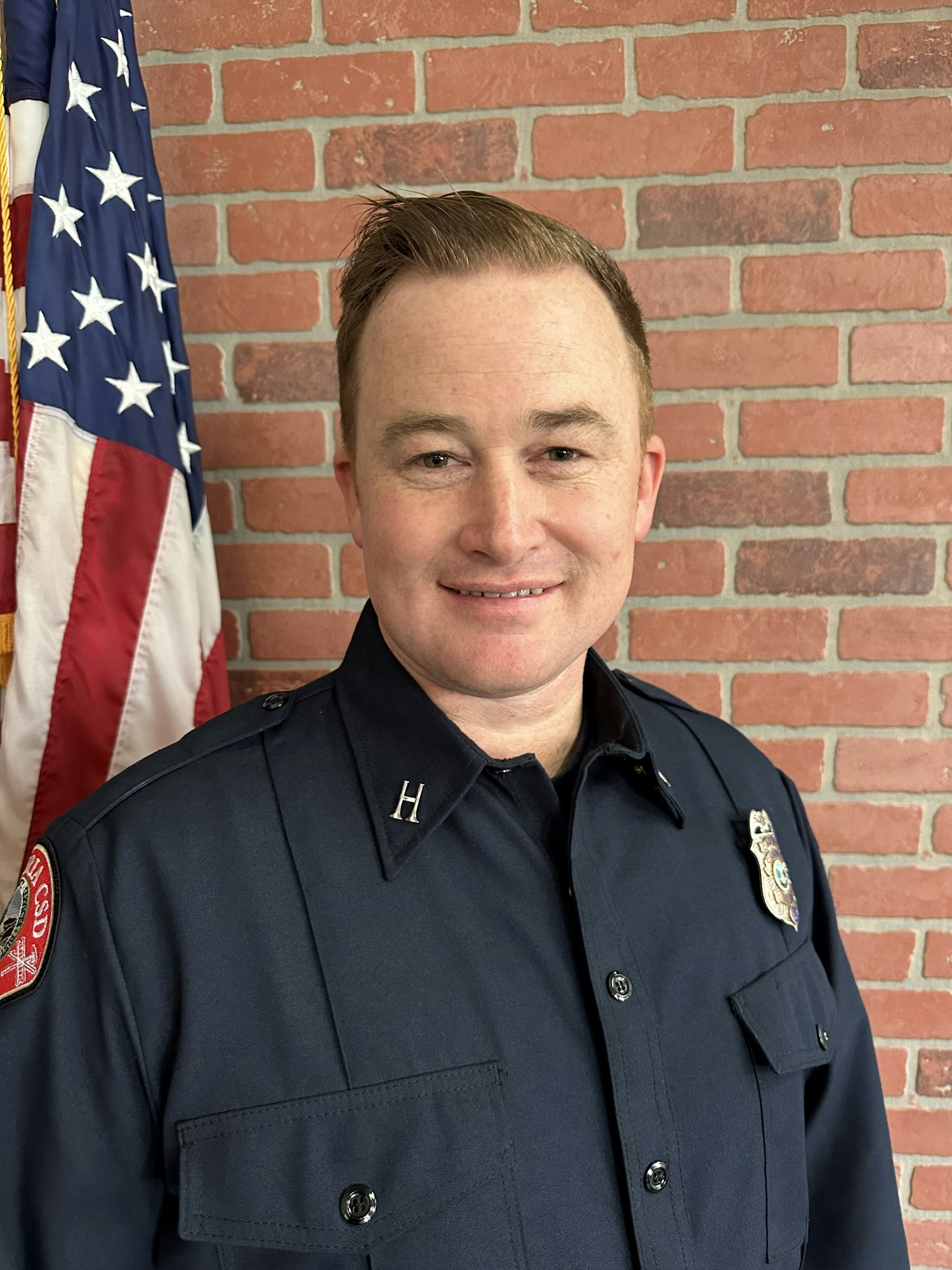A uniformed individual poses in front of a brick wall, with an American flag partially visible beside them.