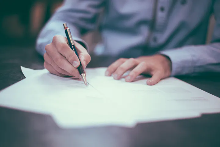 A person writing on sheets of paper, holding a pen, with a close focus on their hands and some documents laid out.
