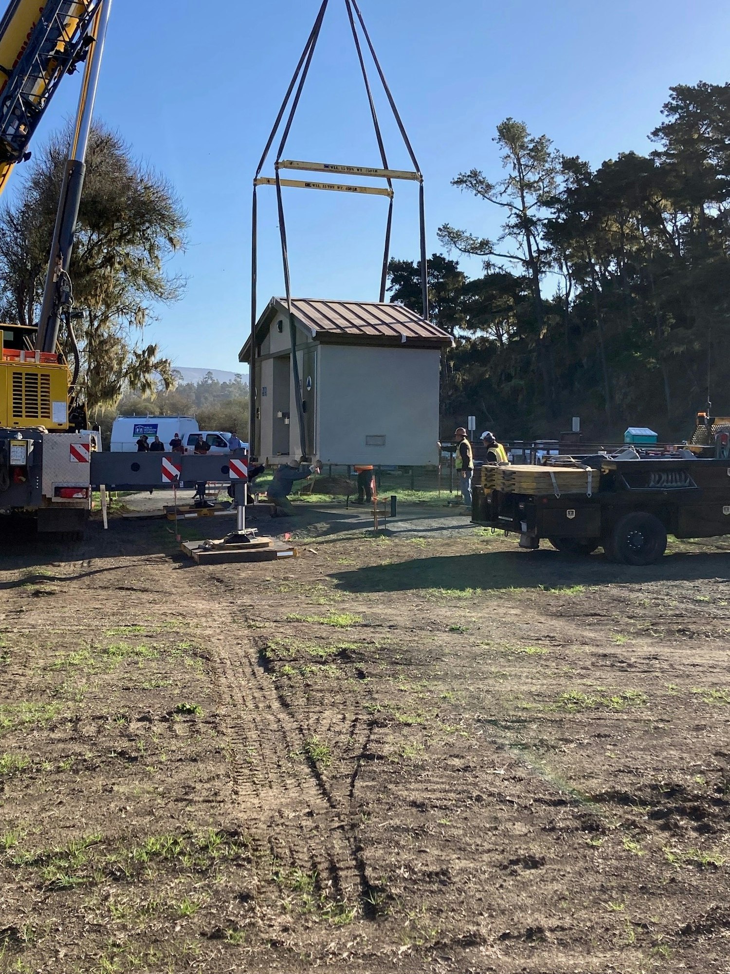 A small building being lifted by a crane with workers and equipment nearby, set against a wooded background.