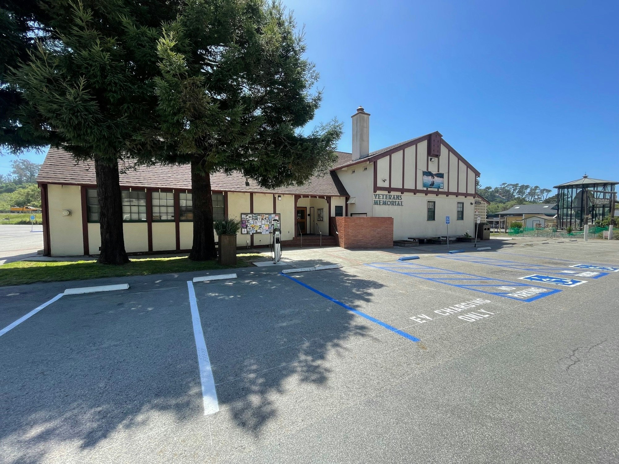 A Tudor-style building with a Veterans Memorial sign, parking lot with handicap spaces, trees, and a clear sky.
