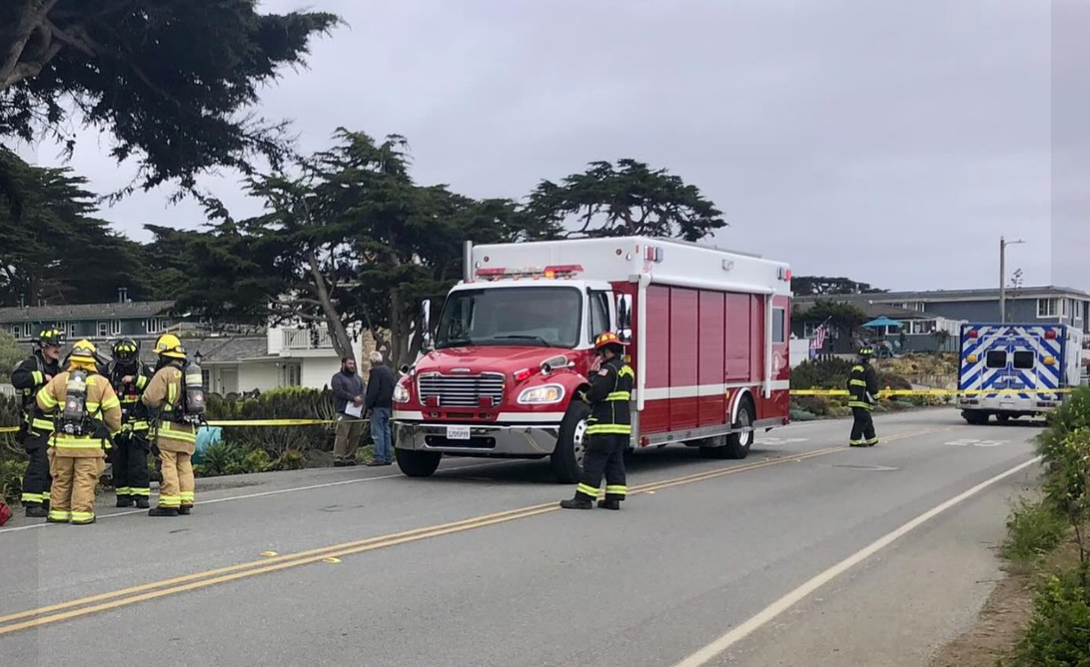 Firefighters near a fire truck and an ambulance at an incident scene, with caution tape and onlookers.