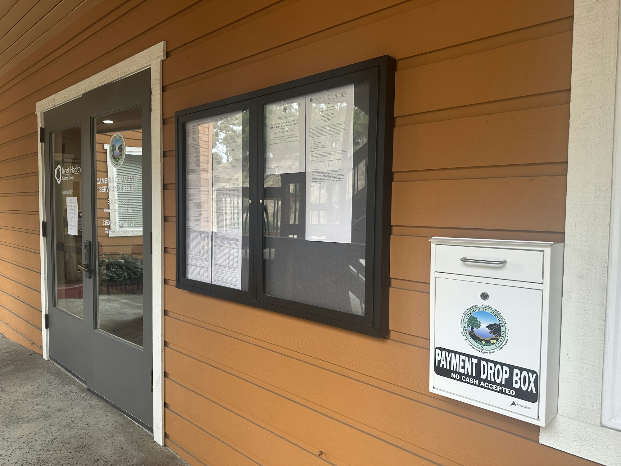 A building entrance with a glass door, notice board, and a "Payment Drop Box" for non-cash payments attached to an orange wall.