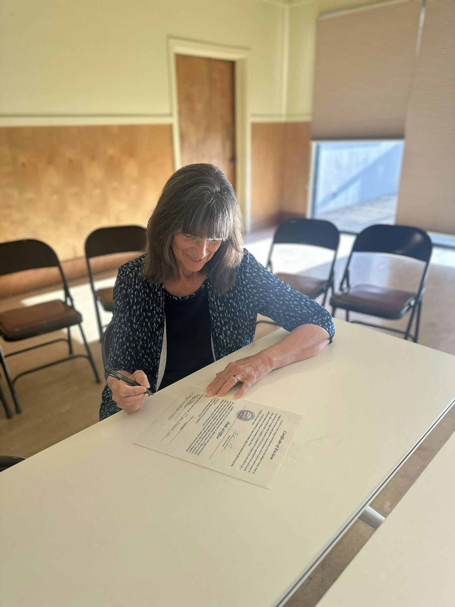A person sits at a table signing a document in a room with chairs.