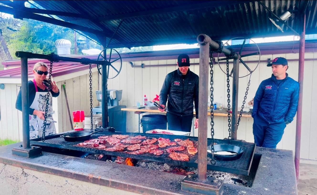 Three people by an outdoor grill with lots of meat cooking over an open fire.