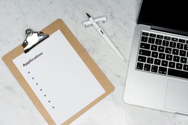 Clipboard with application paper, pen, and laptop on marble surface.