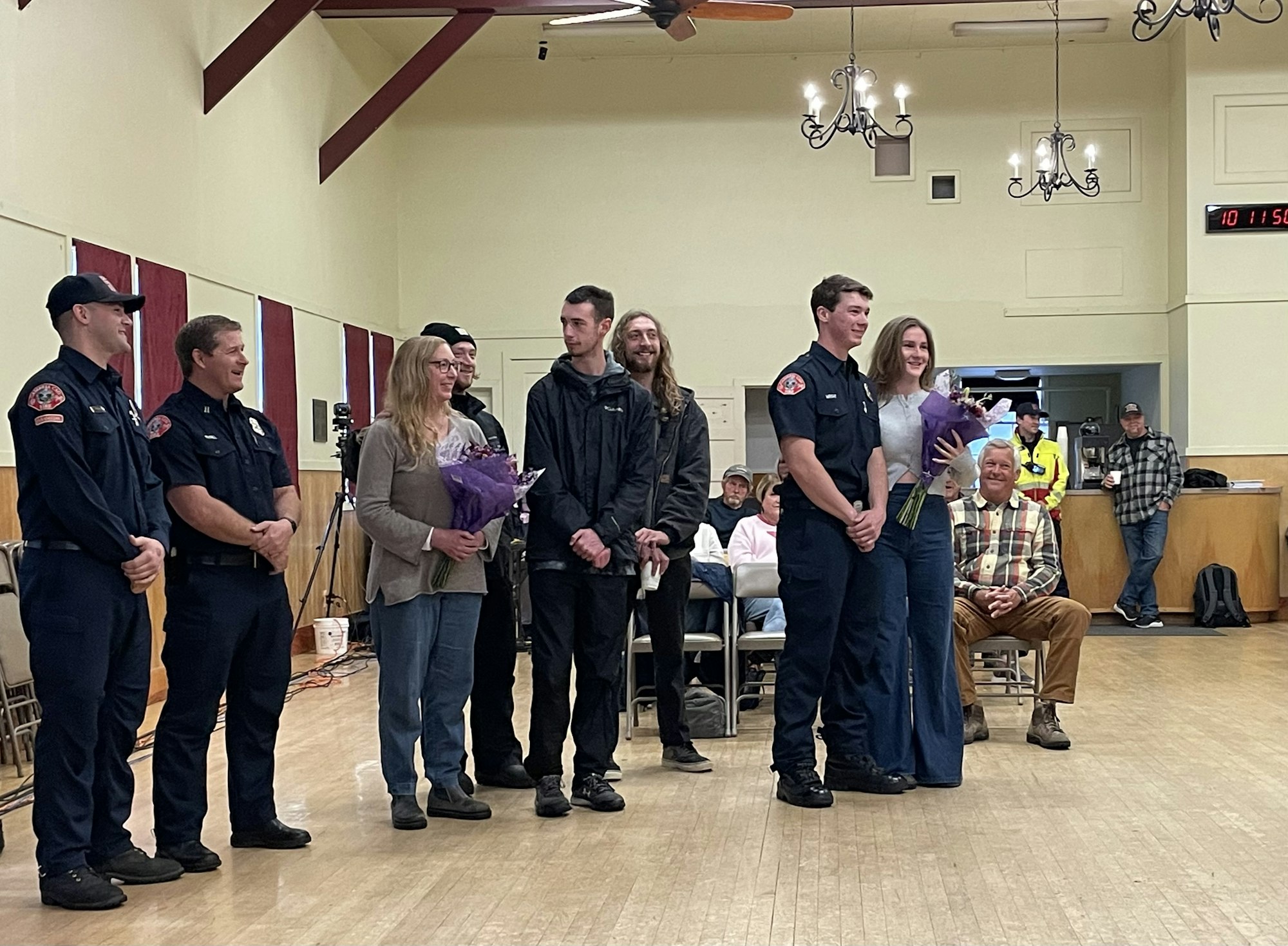 A group of people, some in uniforms, smiling with bouquets, in a hall with onlookers.
