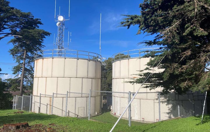 Two large storage tanks with antennas, surrounded by a chain-link fence, against a backdrop of trees and blue sky.