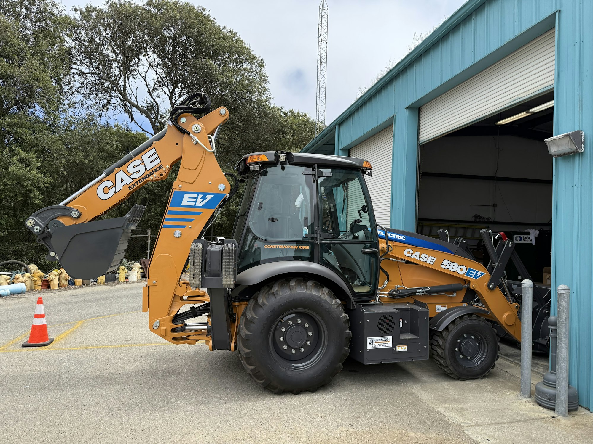A CASE 580 EV backhoe loader next to a building and traffic cone, partially inside the entrance.