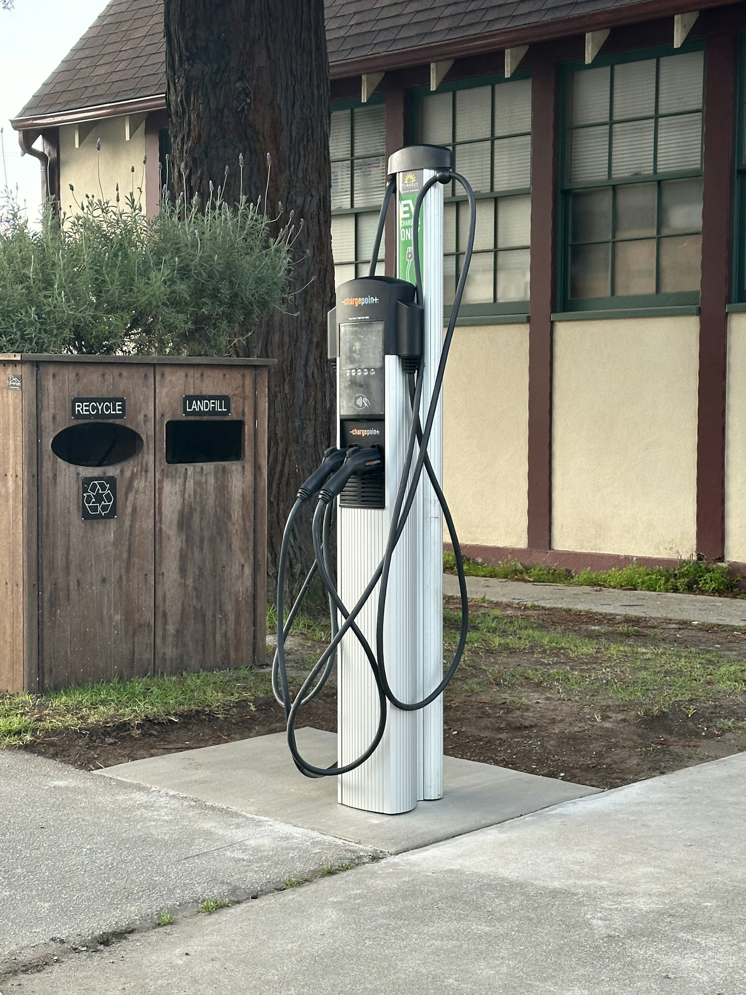 An electric vehicle charging station with cords, next to a trash receptacle, under a shaded structure.