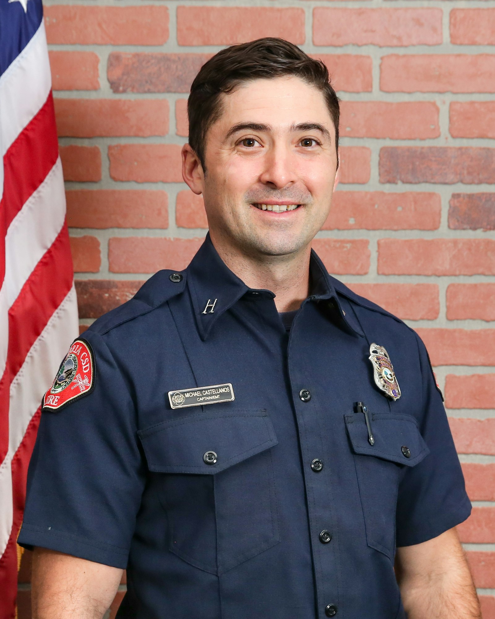 The image features a man in a dark blue firefighter uniform, smiling against a brick background with an American flag.