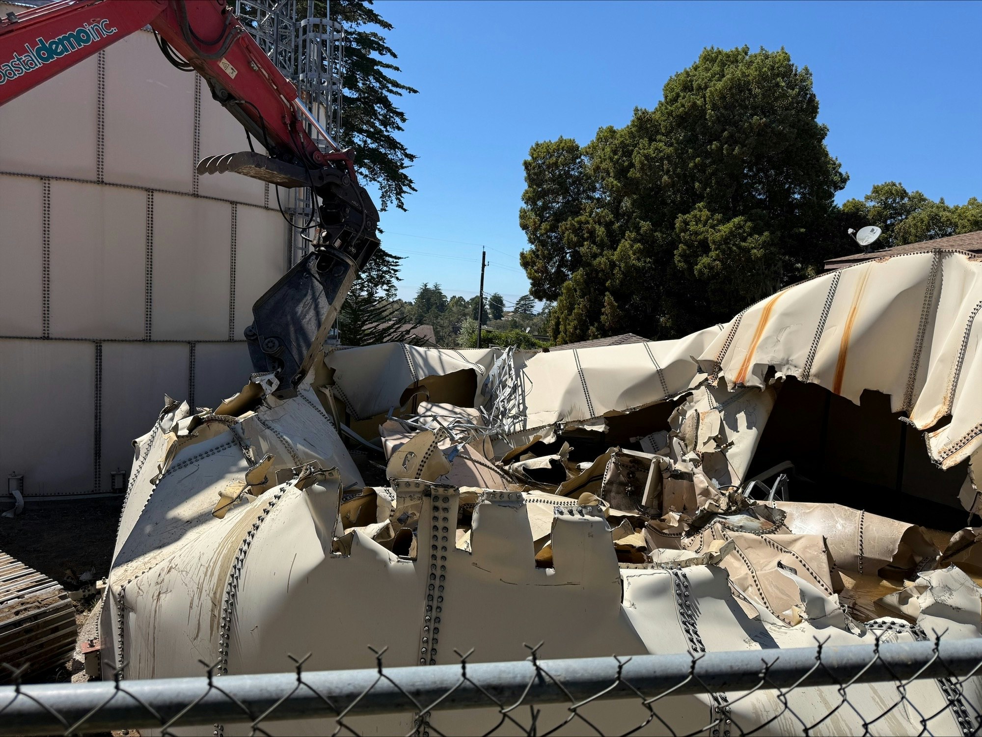 A mechanical claw demolishing a large, collapsed metal structure next to a building, with trees in the background.
