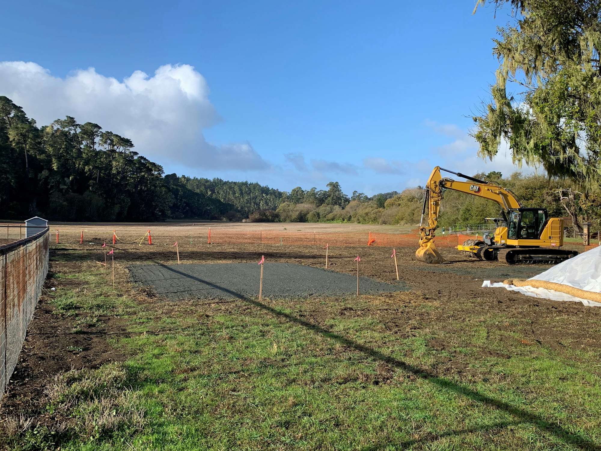 A construction site with an excavator on a grassy field, surrounded by trees and blue sky.