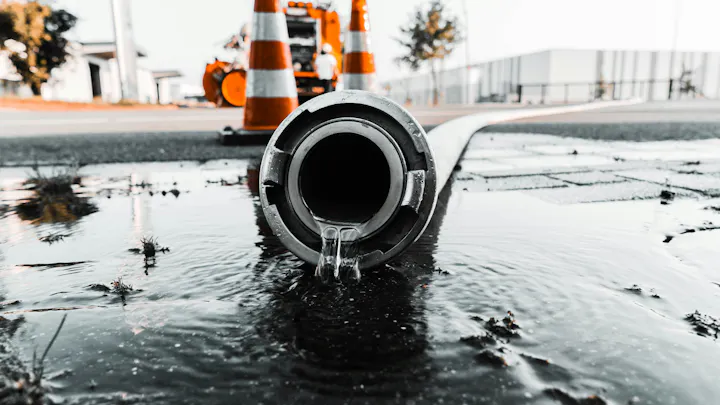 A close-up of a hose pouring water on the ground with traffic cones and construction equipment in the background.