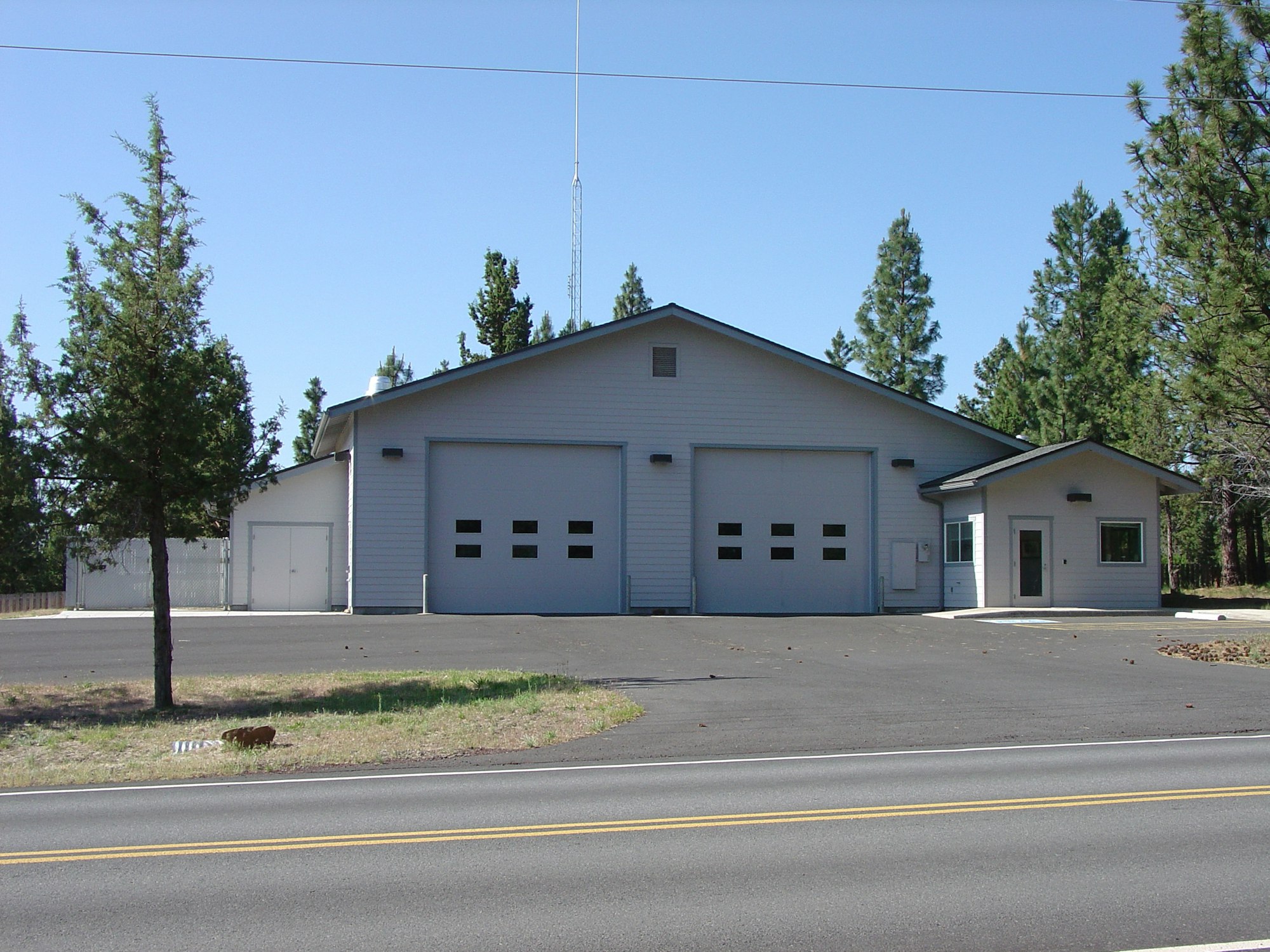 A small fire station with two garage doors, set in a forested area with trees and a clear sky.