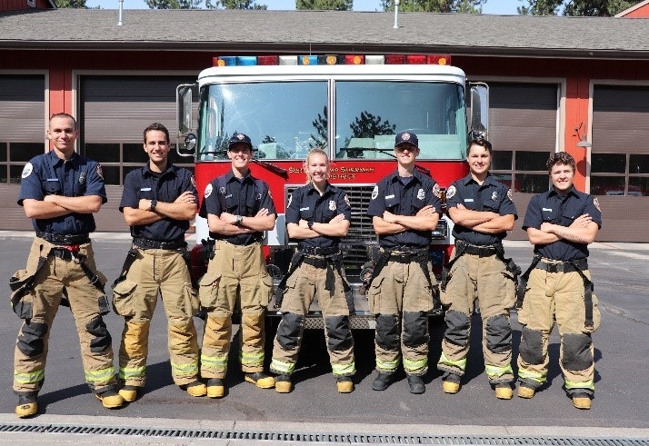 Seven firefighters in uniform stand in front of a fire truck with arms crossed, posing for a group photo.