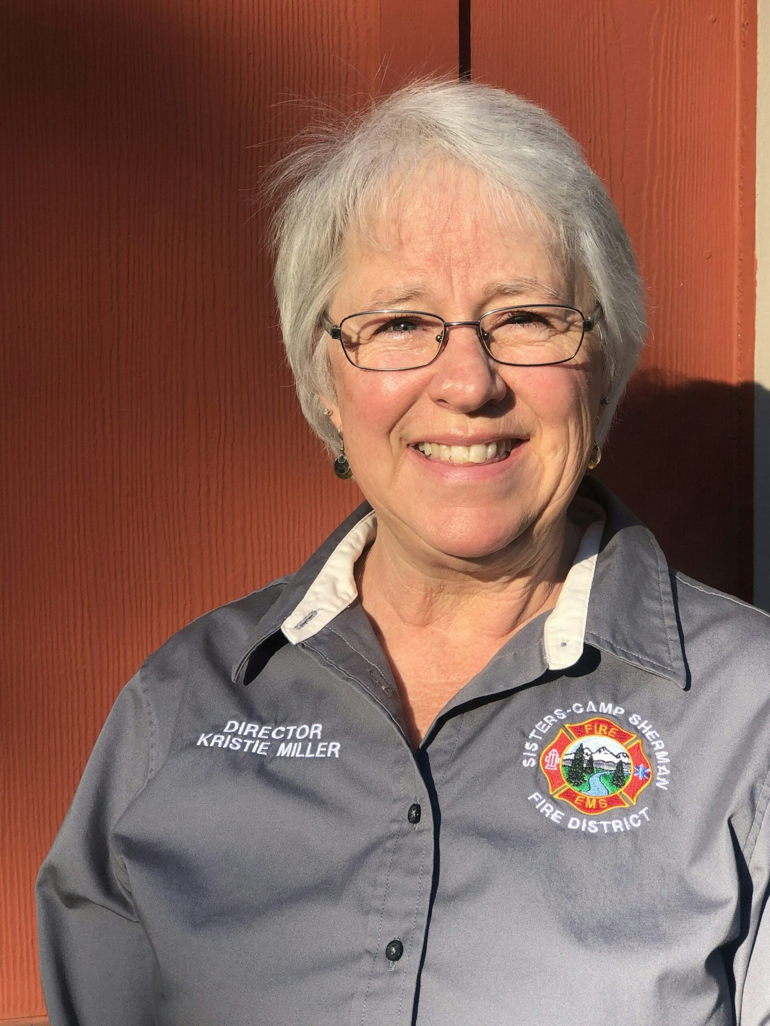 A person in a gray shirt with a fire district patch and "Director Kristie Miller" embroidered on it, standing in front of a red background.