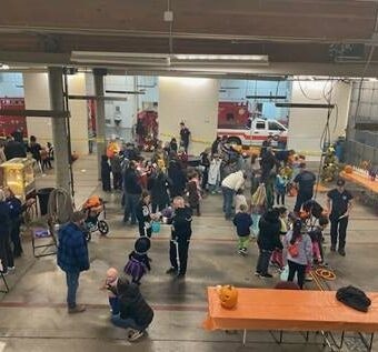 People gathered in a large indoor space with fire trucks, likely a community event. Decorations and pumpkins are visible.