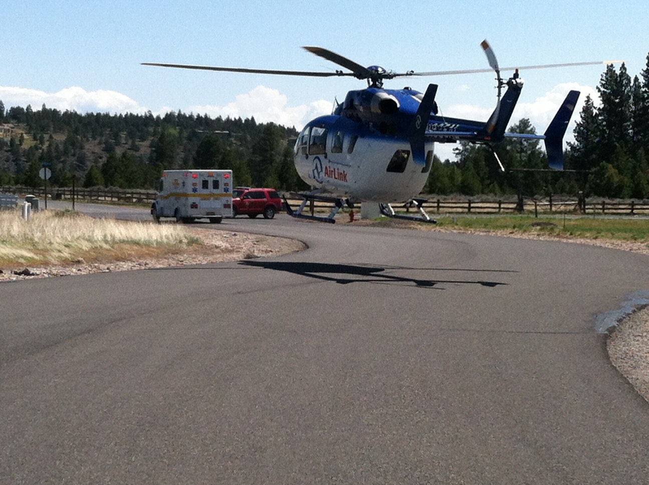 A helicopter landed on a road with an ambulance nearby, surrounded by trees and a fence.