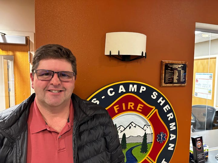 A person in glasses and a jacket stands by a fire department emblem on a wall.