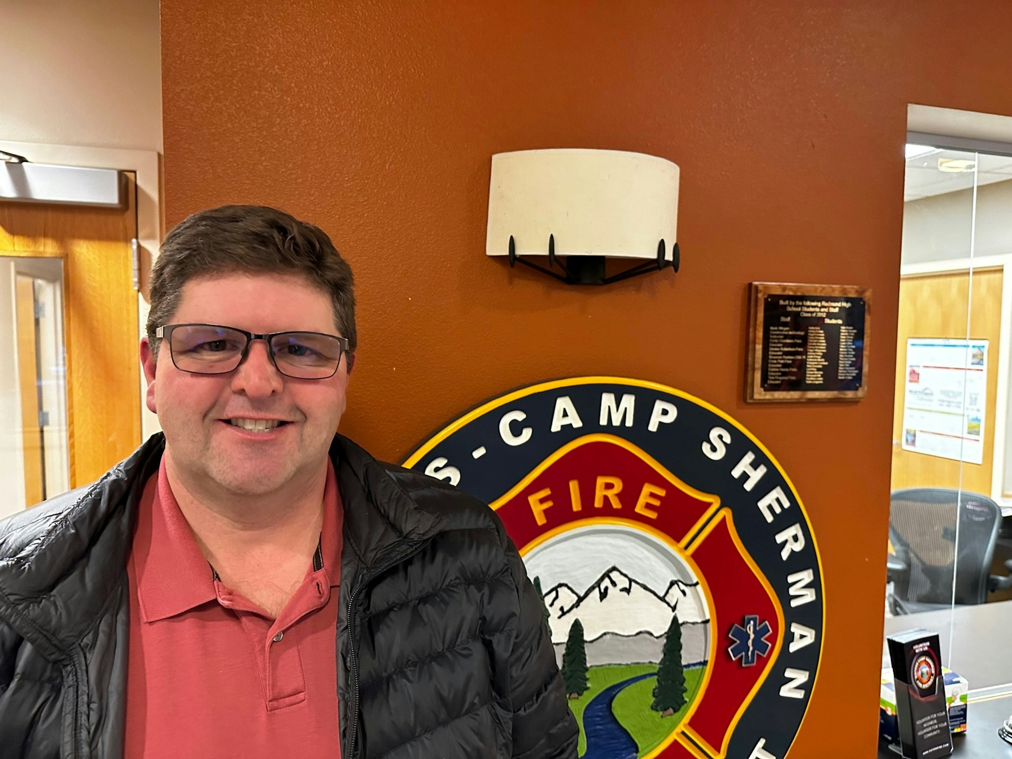 A person in glasses and a jacket stands by a fire department emblem on a wall.