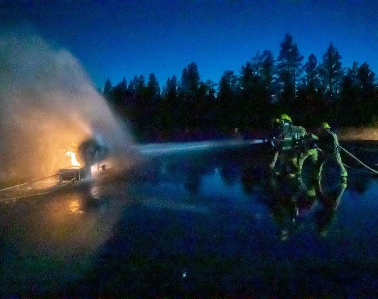 Firefighters extinguishing a blaze at night, using hoses with water spray, against a backdrop of dark trees.