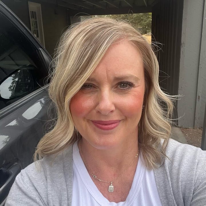 A person with light hair and a necklace, smiling outdoors next to a car.