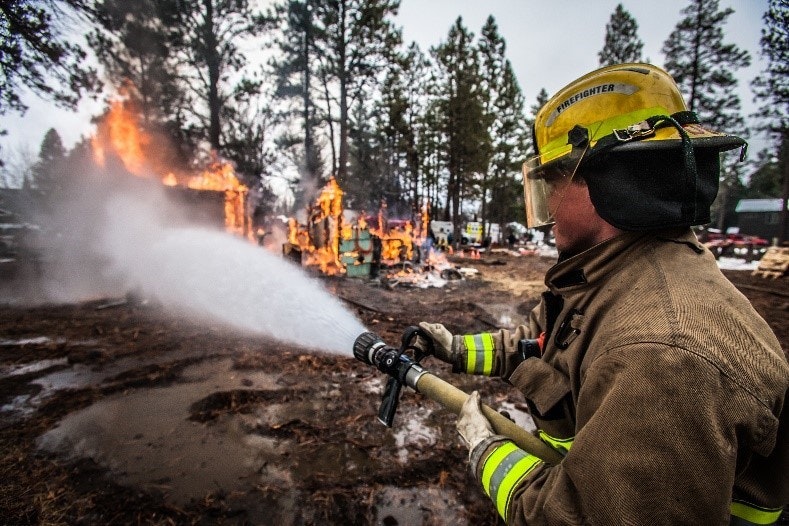 A firefighter sprays water on a burning structure in a forested area.