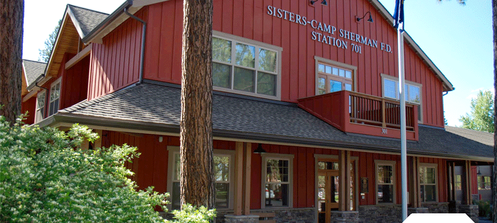 Red building with "SISTERS-CAMP SHERMAN F.D. STATION 701" on it, surrounded by trees and greenery.