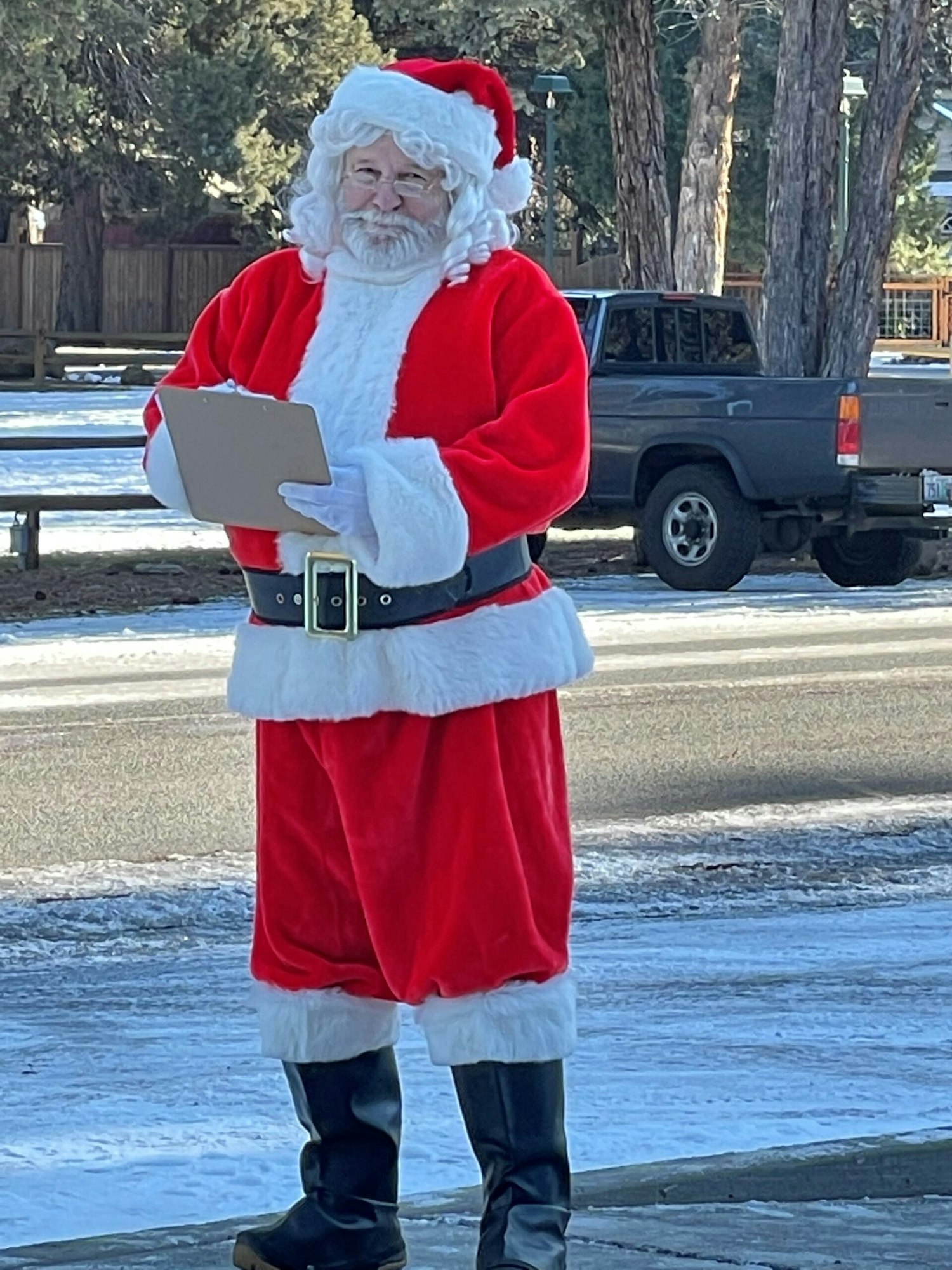 A person dressed as Santa Claus is standing outdoors, holding a clipboard.