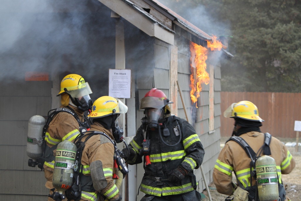 Firefighters in gear near a burning building, smoke and flames visible.