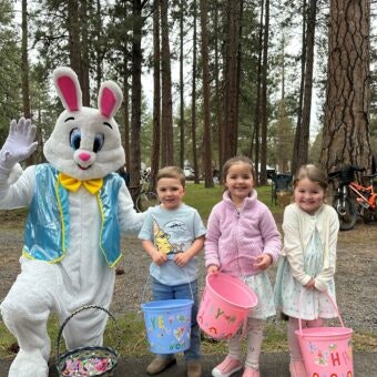 A person in an Easter Bunny costume waves while three children hold colorful buckets in a forested area.
