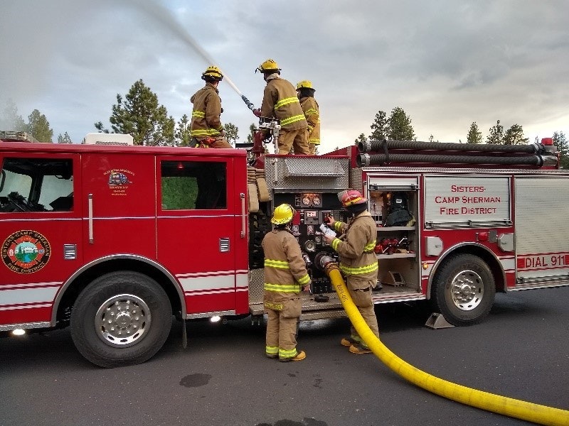 Firefighters in gear operate a red fire truck, with hoses in use, spraying water.