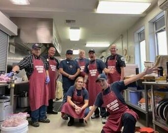 A group of people in a kitchen wearing red aprons, some kneeling, others standing. They appear to be posing for a group photo.