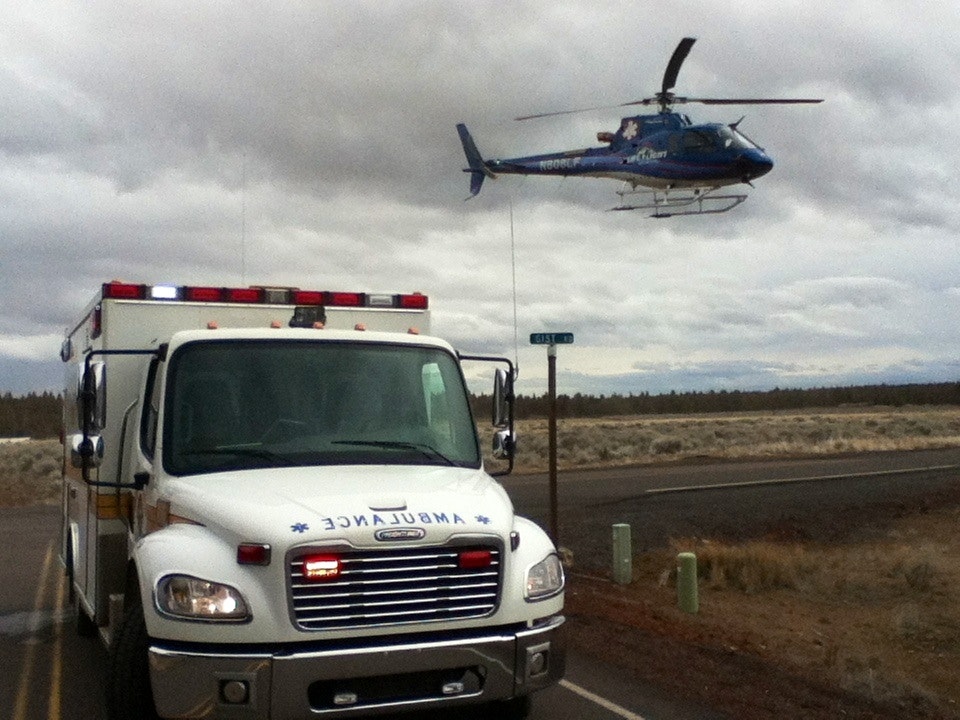 An ambulance is parked on a road while a helicopter hovers above in a rural area.