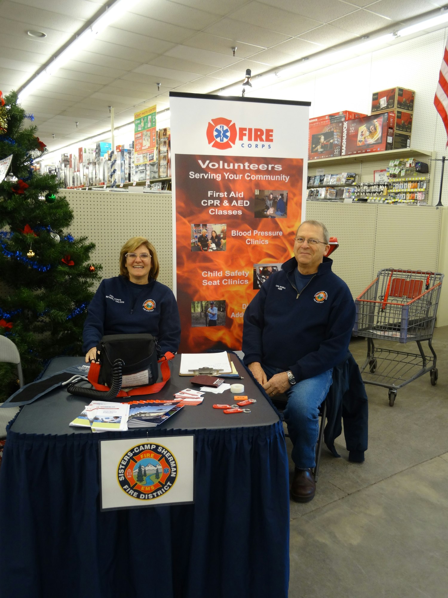 Two people at a "Fire Corps" volunteer recruitment table with brochures in a store.