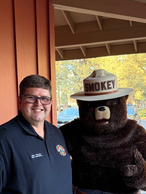 A man in a shirt smiles next to a bear mascot wearing a hat labeled "SMOKEY." They are outside with trees in the background.