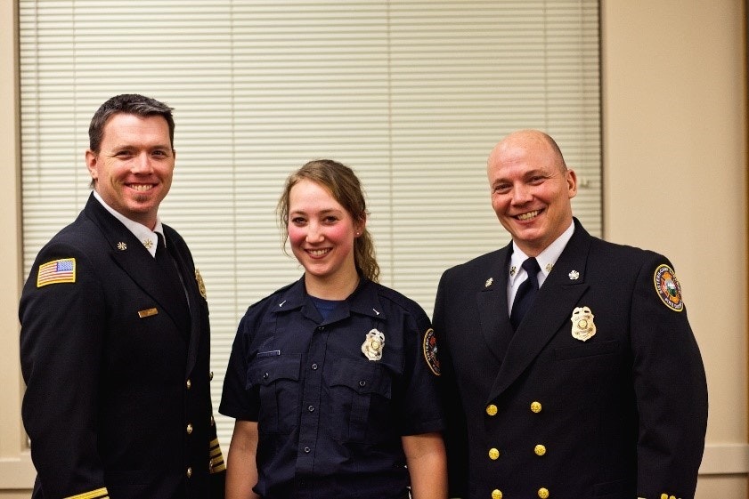 Three people in uniforms, two men and one woman, possibly firefighters, smiling indoors.