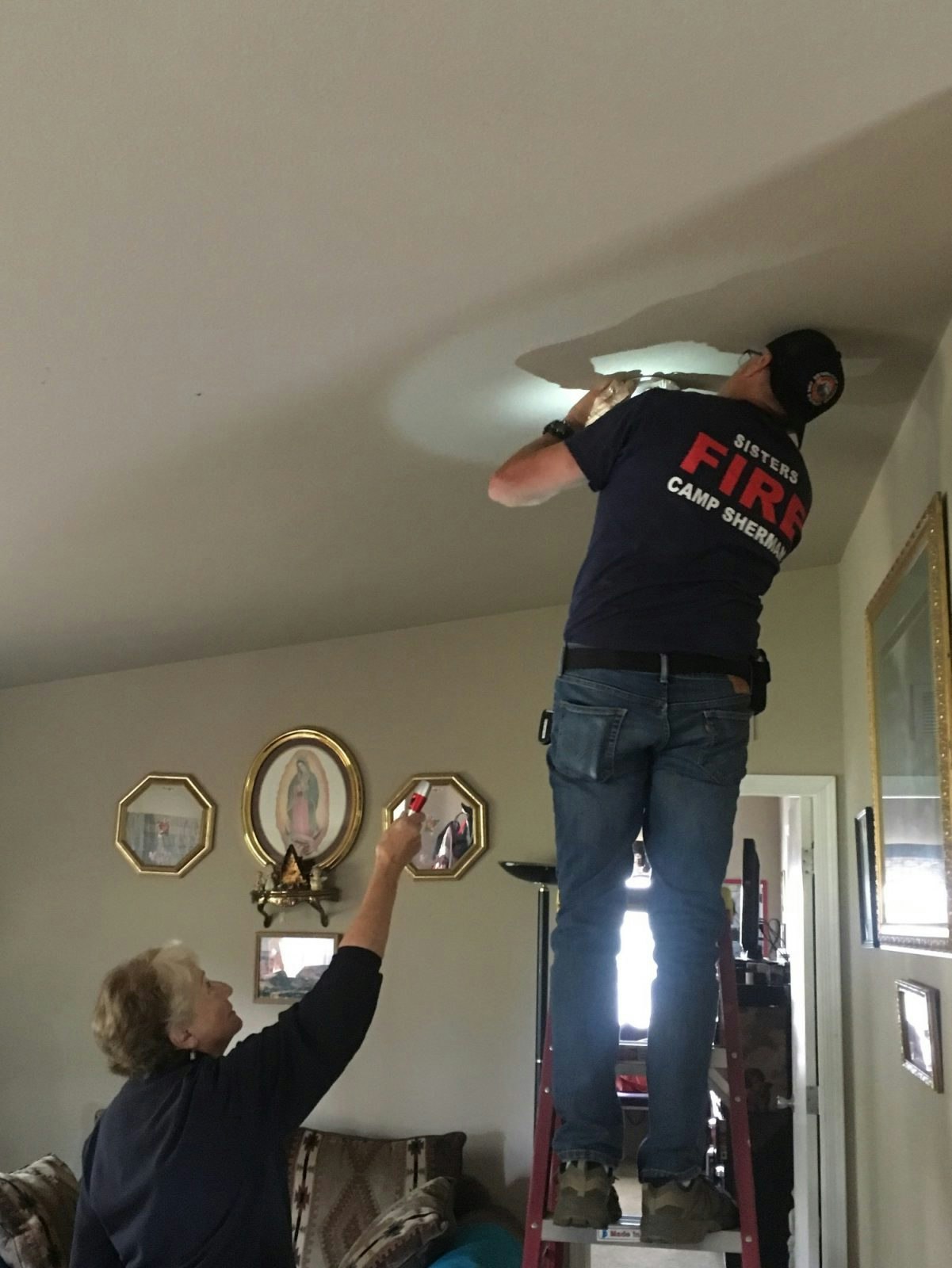 A firefighter on a ladder inspects a ceiling while a woman holds up a flashlight.