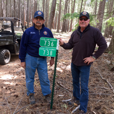 Two people in a forest stand beside a signpost labeled "Sisters-Camp Sherman 736, 738." A vehicle is nearby.
