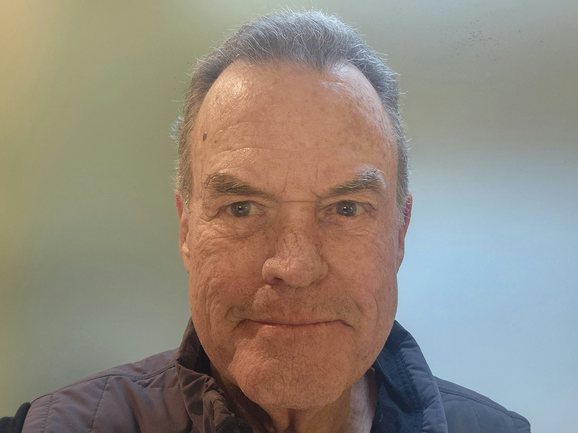 The image features an older man with short hair, wearing a vest, smiling against a neutral background.