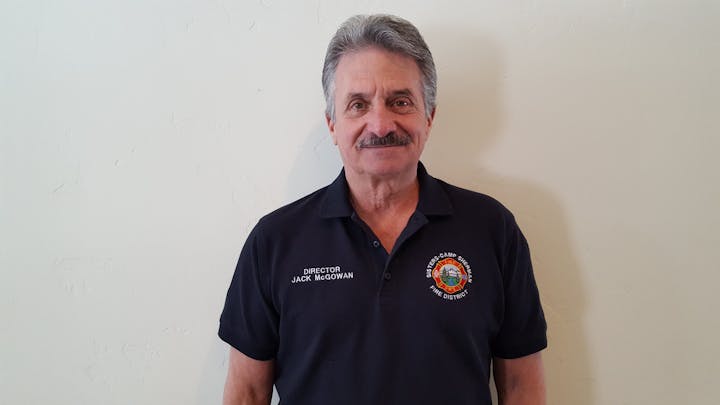 A man in a navy polo shirt with a fire department logo and text "Director Jack McGowan" stands against a plain wall.